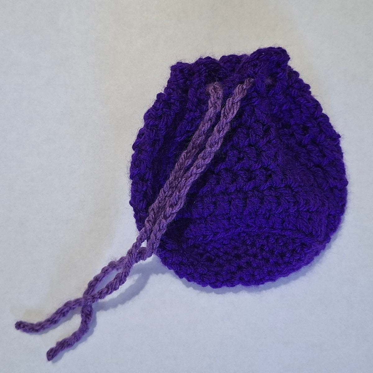 A small handmade purple crocheted drawstring pouch or bag, photographed on a plain off-white background.